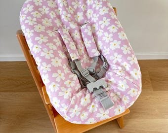May include: A baby high chair cushion with a pink background and white daisy pattern. The cushion has a gray safety harness and is placed on a wooden high chair. The cushion is designed to provide comfort and support for infants and toddlers.