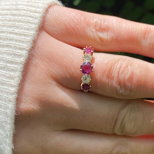 May include: A gold band ring with alternating pink and white gemstones. The ring is set with round stones and has a polished finish.