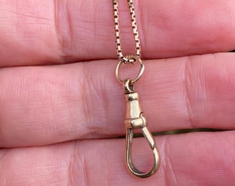 May include: A gold-colored necklace with a box chain and a clasp. The clasp is a lobster claw style. The necklace is held in a person's hand.