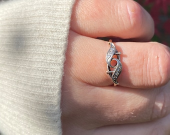 May include: A gold ring with a delicate design featuring two curved lines set with small diamonds. The ring is on a person's finger.