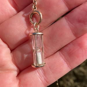 May include: A gold-toned metal hourglass pendant featuring a clear glass chamber filled with fine white sand. The miniature hourglass is attached to a circular gold-toned metal spring ring clasp, designed to be worn as a charm on a necklace or bracelet.