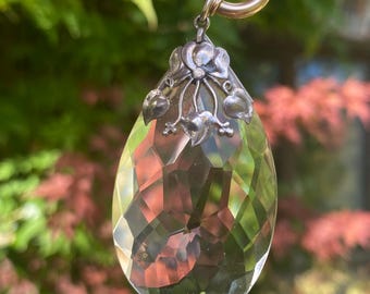 May include: A faceted, teardrop-shaped pendant with a silver floral design and a gold-colored ring. The clear crystal reflects light, creating a sparkling effect. The pendant is suspended against a blurred green and red background.