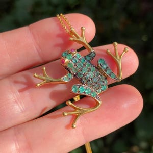 Puede incluir: Colgante de rana dorada con detalles en verde esmeralda y negro. Los ojos de la rana son pequeñas gemas rojas. El colgante está sujeto a una delicada cadena dorada. Las patas de la rana están extendidas.