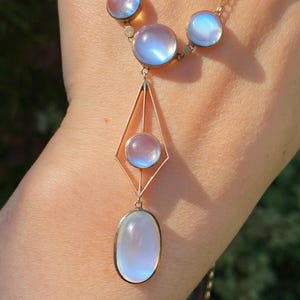 May include: A gold-toned necklace featuring iridescent, round and oval moonstone pendants. The necklace has a geometric design with a diamond-shaped element and a delicate chain. The moonstones display a soft, ethereal glow, reflecting shades of blue and white.
