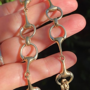 May include: A gold-toned metal bracelet with a chain link fastening. The bracelet is made of a series of interlocking horse bit-shaped links.