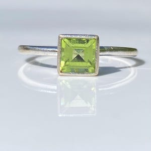 May include: A silver ring with a square, light green peridot gemstone. The gemstone is set in a square bezel and has a faceted appearance. The ring band is thin and polished, reflecting light. The ring is displayed on a white surface.