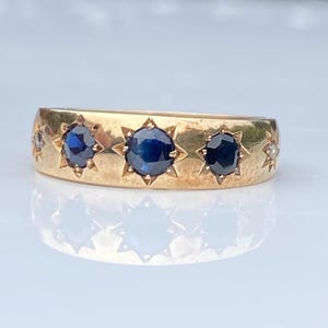 May include: A gold band ring featuring three prominent round blue sapphires set in star-shaped engravings across the center, flanked by two smaller clear stones on the sides, all embedded within the smooth, polished surface of the metal band.
