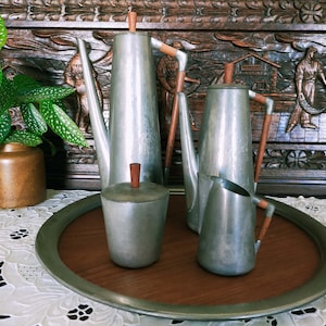 Peut inclure: Un ensemble de trois théières en métal argenté avec des poignées en bois sur un plateau en bois. Les théières sont sur une nappe en dentelle blanche.