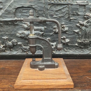 May include: A vintage metal press, likely for crafts or small manufacturing, mounted on a wooden base. The press has a lever, a screw mechanism, and a detailed, ornate design. The background features a dark, textured relief sculpture.