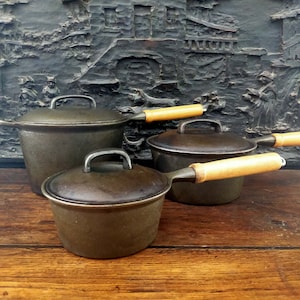 May include: Three cast iron pots with wooden handles. The pots are nested together, with the largest pot at the bottom and the smallest pot on top. The pots are all dark grey in colour and have a rustic appearance.