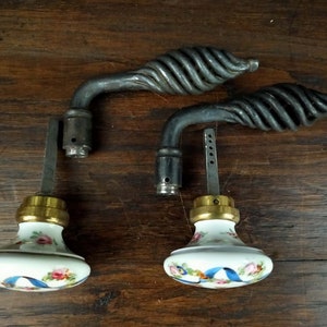 May include: Two antique door handles with porcelain knobs. The handles are made of twisted metal and have a black finish. The knobs are white with a floral design and blue ribbon accents.