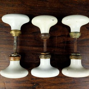 May include: Three white porcelain door knobs with brass hardware components. Each knob features a smooth, rounded white handle connected to a central metal spindle and a circular brass base plate, showing signs of age and patina on the metal parts.