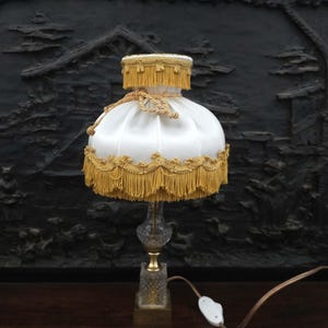 Könnte beinhalten: Eine Vintage-Tischlampe mit einem weißen Seidenschirm, der mit goldenen Fransen und Zierleisten verziert ist. Die Lampe hat einen Sockel aus klarem Glas mit einem goldfarbenen Metallakzent und einem dekorativen quadratischen Sockel. Die Lampe ist eingesteckt und betriebsbereit.