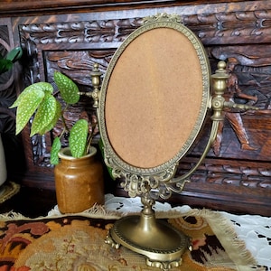 May include: A vintage brass oval mirror with ornate detailing and a stand. The mirror is on a table with a patterned fabric and a white lace doily.