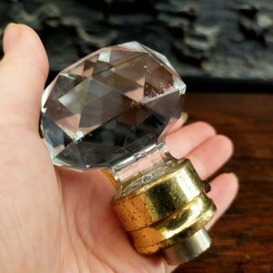 Peut inclure: Un bouton en verre transparent à facettes en forme de diamant avec une base en métal doré poli. Cette pièce de quincaillerie décorative présente un sommet à facettes multiples qui reflète la lumière, fixé à une douille cylindrique en métal doré.