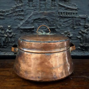 May include: A copper pot with a lid and two side handles. The pot has a rounded body and a small handle on the lid. The pot is sitting on a wooden surface. The background is a dark, textured wall.