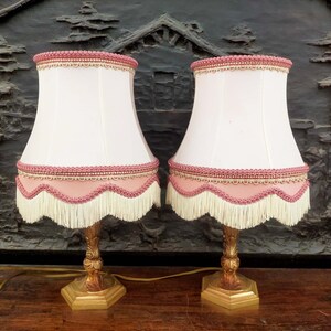 Peut inclure: Deux lampes de table dorées avec des abat-jours blancs bordés de franges roses et d'une bande de perles roses.
