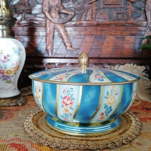 Puede incluir: Una caja de hojalata vintage azul y blanca con un diseño floral y ribetes dorados. La caja tiene una tapa y está decorada con rayas y flores.