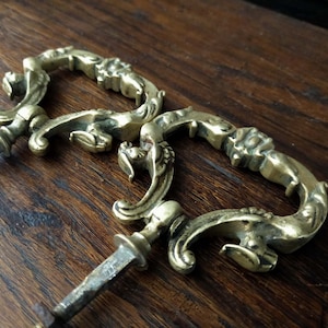 Könnte beinhalten: Zwei verzierte Messing-Schubladengriffe mit herzförmigem Design. Die Griffe haben ein detailliertes, rollenförmiges Muster und ein spitzes Ende.