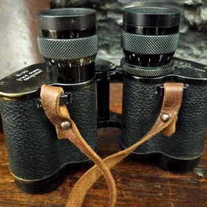 Può includere: Binocolo nero con cinturino in pelle marrone. Il binocolo ha una finitura nera testurizzata e una fascia in ottone attorno al corpo. Il cinturino ha una fibbia in metallo.