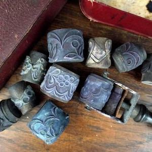 Pode incluir: Uma coleção de blocos de impressão de madeira vintage com designs intrincados. Os blocos estão em vários tons de azul e roxo, e alguns têm um aspecto desgastado e antigo. Os blocos estão dispostos sobre uma superfície de madeira, e há uma pequena prensa de impressão de metal no fundo.