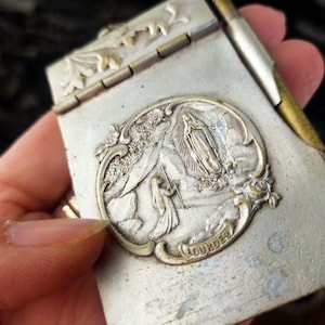 Könnte beinhalten: Eine Vintage-Metallbox in Silberoptik mit Klappdeckel. Die Box hat ein geprägtes ovales Medaillon mit einer religiösen Szene und dem Wort "Lourdes". Die Box wird in einer Hand gehalten.