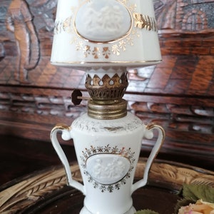 Op de afbeelding: Een witte porseleinen olielamp met gouden accenten en een witte kap. De lamp heeft een reliëfdesign van een stel in een romantische omhelzing. De kap heeft een gouden rand en een bloemenpatroon.