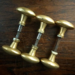 Peut inclure: Trois poignées de porte anciennes en laiton de forme ovale, disposées sur une surface en bois foncé. Les poignées ont un design classique, avec une base cylindrique et une longue tige métallique.