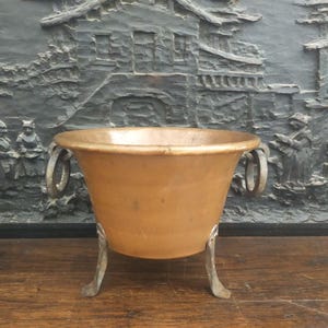 Peut inclure: Un bol en métal de couleur cuivre avec deux poignées et trois pieds de couleur argent. Le bol a une large ouverture et un corps légèrement conique. Le fond présente une sculpture en relief sombre et texturée représentant une scène de village.