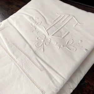 May include: Folded white linen textile with embroidered floral and initial details. The embroidery includes a large initial, floral sprigs, and decorative stitching. The fabric is folded, showing multiple layers.
