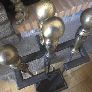 Może przedstawiać: Zestaw narzędzi do kominka w stylu vintage z czarnym metalowym stojakiem. Zestaw zawiera pogrzebacz, łopatę i szczotkę, każdy z mosiężnymi uchwytami w kształcie kuli. Narzędzia są ustawione przed ceglanym kominkiem.