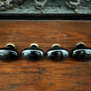 Op de afbeelding: Vier zwarte ovale kastknoppen met messingkleurige bases liggen op een houten oppervlak. De knoppen hebben een glanzende afwerking, die de omgeving weerspiegelt. Het hout heeft een warme, bruine tint. Geschikt voor meubels.
