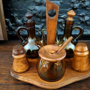 Puede incluir: Un juego de condimentos de madera con cuatro botellas de vidrio y una cuchara de madera. Las botellas son de vidrio marrón y tienen tapones de madera. El juego está sobre una base de madera.