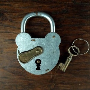 Peut inclure: Un cadenas en argent avec un trou de serrure et un levier en laiton. Le cadenas a une forme arrondie avec deux petites oreilles sur le dessus. La clé est en laiton et a un anneau à l'extrémité.