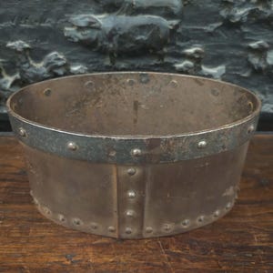 Peut inclure: Un bol en métal riveté avec un bord renforcé, montrant des signes d'âge. Le bol a une forme cylindrique avec un dessus légèrement évasé. Le métal a un aspect terni, suggérant qu'il s'agit d'une pièce ancienne ou vintage. Il repose sur une surface en bois.