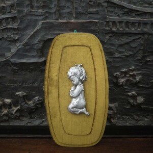 Peut inclure: Un bas-relief argenté d'un enfant agenouillé en prière est monté sur une plaque ovale jaune moutarde. La plaque a un aspect texturé, semblable à du velours côtelé. Le fond est un mur sombre et texturé.