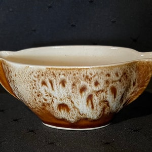 May include: A ceramic serving bowl with a distinctive design. The bowl has a cream-coloured interior and a textured exterior with a brown and white speckled pattern. The bowl has a curved shape with a spout on each side.