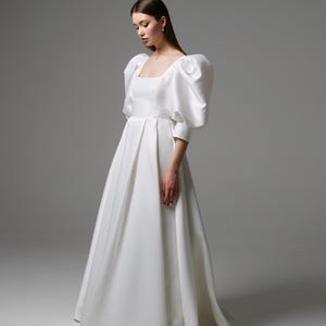 May include: A floor-length white gown featuring a square neckline, a fitted bodice, and voluminous puff sleeves that extend to the elbows. The skirt is full and pleated, falling from the waist to the floor in a smooth, structured fabric.