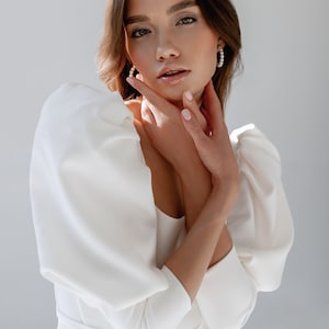 May include: A woman wearing a white dress with large, puffy sleeves. She is looking at the camera with her hands on her neck.