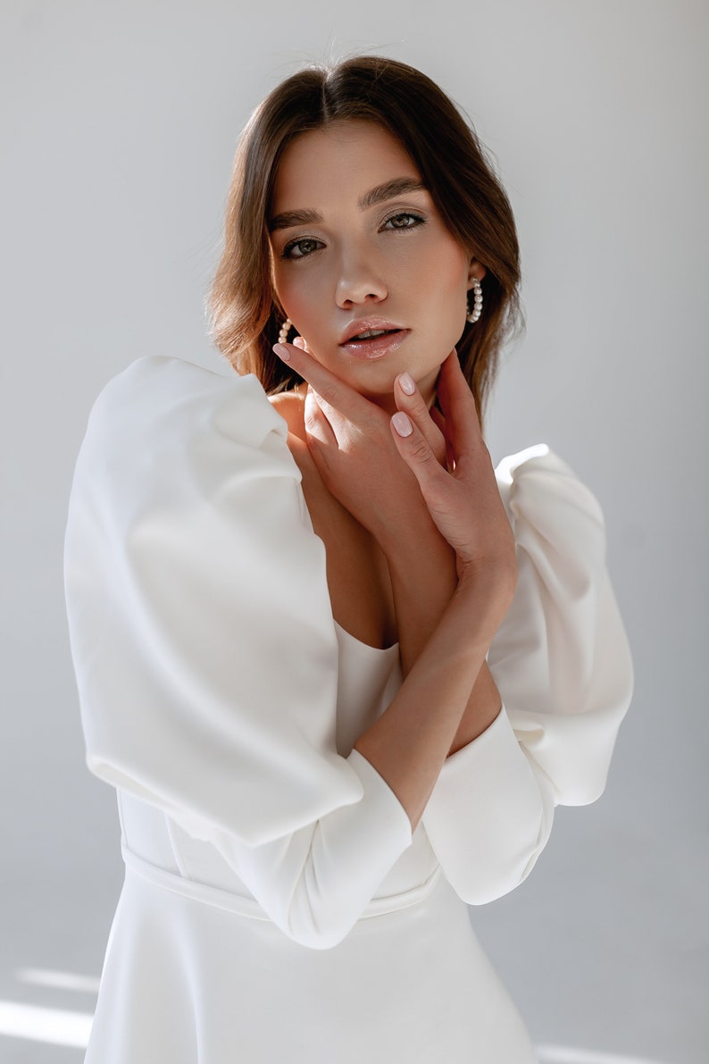 May include: A woman wearing a white dress with large, puffy sleeves. She is looking at the camera with her hands on her neck.