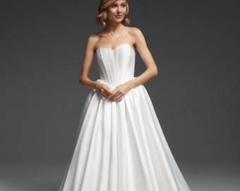 May include: A strapless white wedding gown with a fitted bodice and a full, flowing skirt. The dress features vertical pleats on the bodice and a classic, elegant design. The model is wearing a necklace.