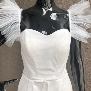 K&ouml;nnte beinhalten: Wei&szlig;er Festtags-Jumpsuit mit herzf&ouml;rmigem Ausschnitt und zarter Schleife in der Taille. Der Jumpsuit hat transparente, ger&uuml;schte &Auml;rmel. Der Stoff hat eine subtile Textur, die einen raffinierten Look erzeugt.