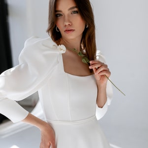 May include: A woman wearing a white, long-sleeved dress with a square neckline and puffed sleeves. The dress has a fitted bodice and a flowing skirt. She is holding a single white flower.