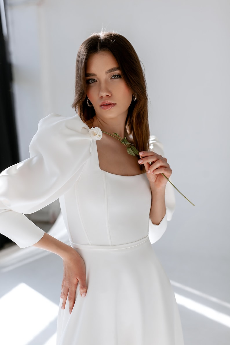 May include: A woman wearing a white, long-sleeved dress with a square neckline and puffed sleeves. The dress has a fitted bodice and a flowing skirt. She is holding a single white flower.