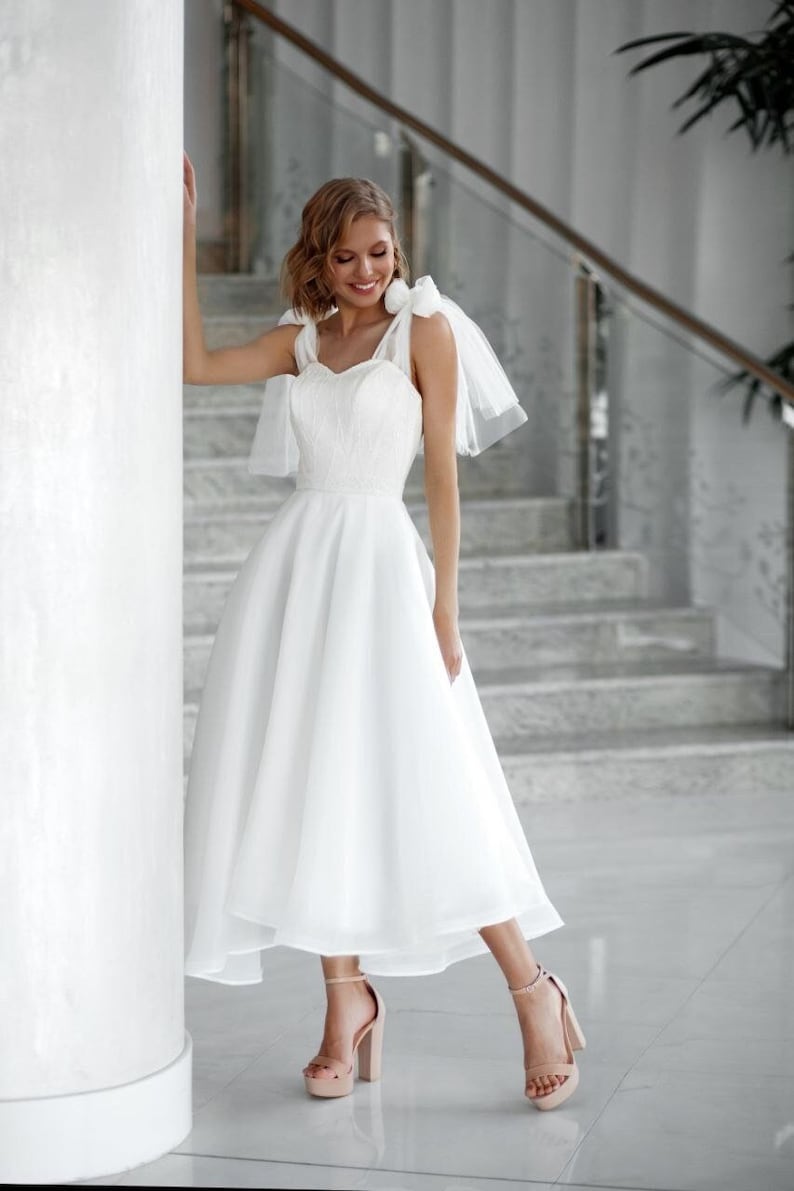 May include: A white wedding dress with a sweetheart neckline and a flowing skirt. The dress features delicate detailing on the bodice and is paired with nude platform heels. The bride is smiling, leaning against a white column.