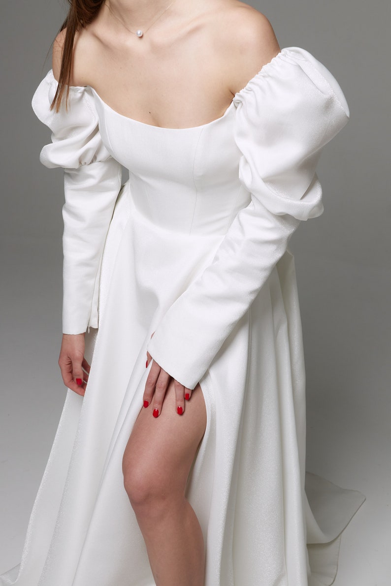 Puede incluir: Un vestido blanco de hombros descubiertos con mangas largas y abullonadas y una abertura en la pierna hasta el muslo. El vestido tiene un corpi&ntilde;o ajustado y una falda fluida.