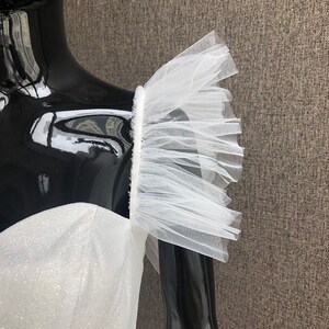 K&ouml;nnte beinhalten: Ein wei&szlig;es, paillettenbesetztes Oberteil mit einem T&uuml;llr&uuml;schen&auml;rmel-Detail. Das Kleid wird auf einer schwarzen Schaufensterpuppe pr&auml;sentiert, die die zarte Textur und das Design der Abendgarderobe zeigt.