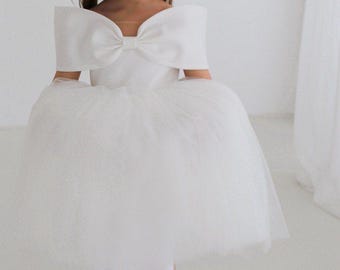 Abito da damigella in raso bianco, gonna a balze in tulle, fiocco grande, maniche a spalle scoperte.