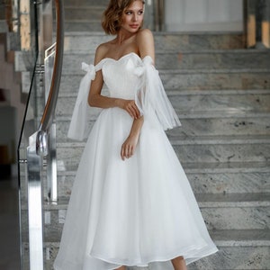 May include: A white, off-the-shoulder wedding dress with a sweetheart neckline and a knee-length skirt. The dress features sheer, flowing sleeves tied with bows at the shoulders. The model is wearing nude platform heels and standing on a marble staircase.