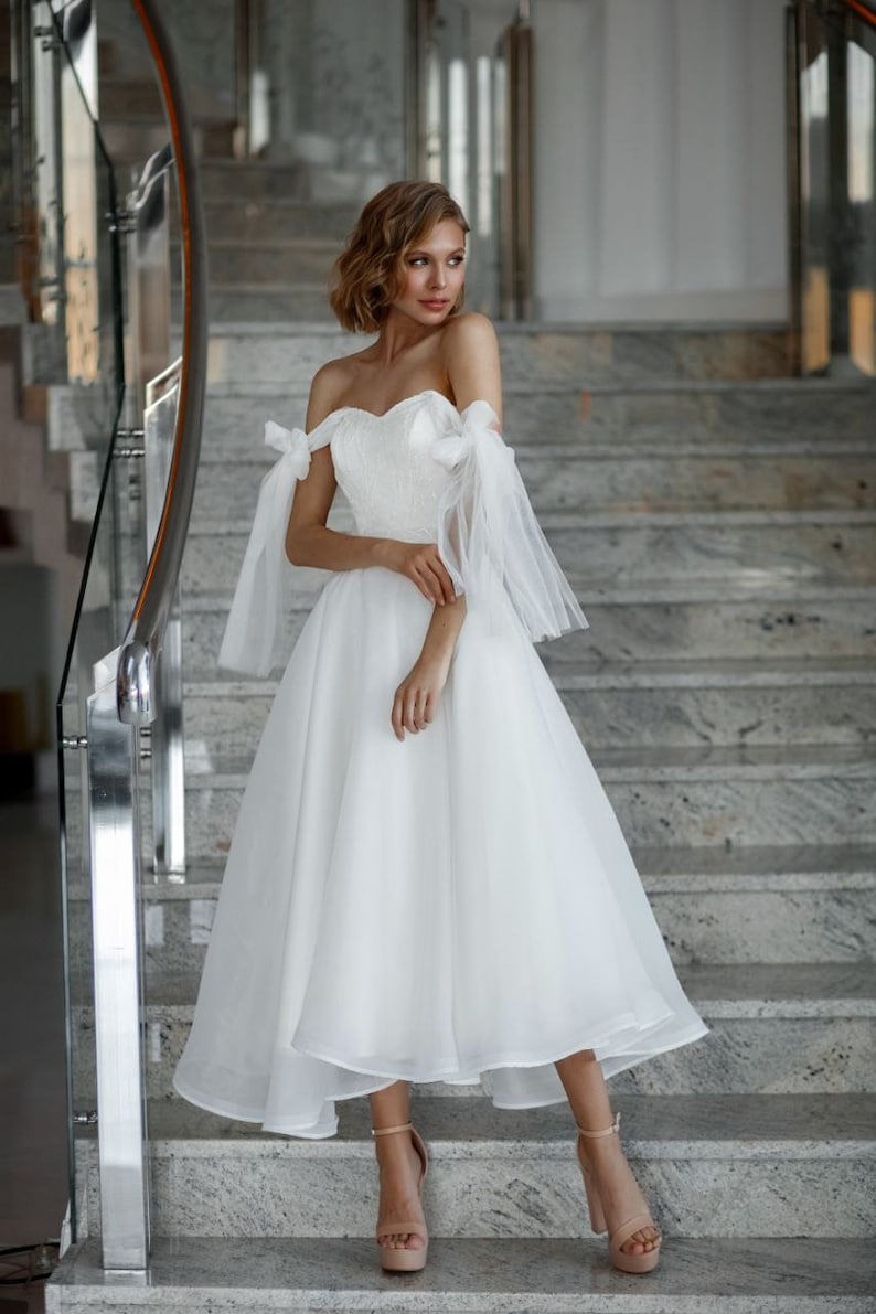 May include: A white, off-the-shoulder wedding dress with a sweetheart neckline and a knee-length skirt. The dress features sheer, flowing sleeves tied with bows at the shoulders. The model is wearing nude platform heels and standing on a marble staircase.